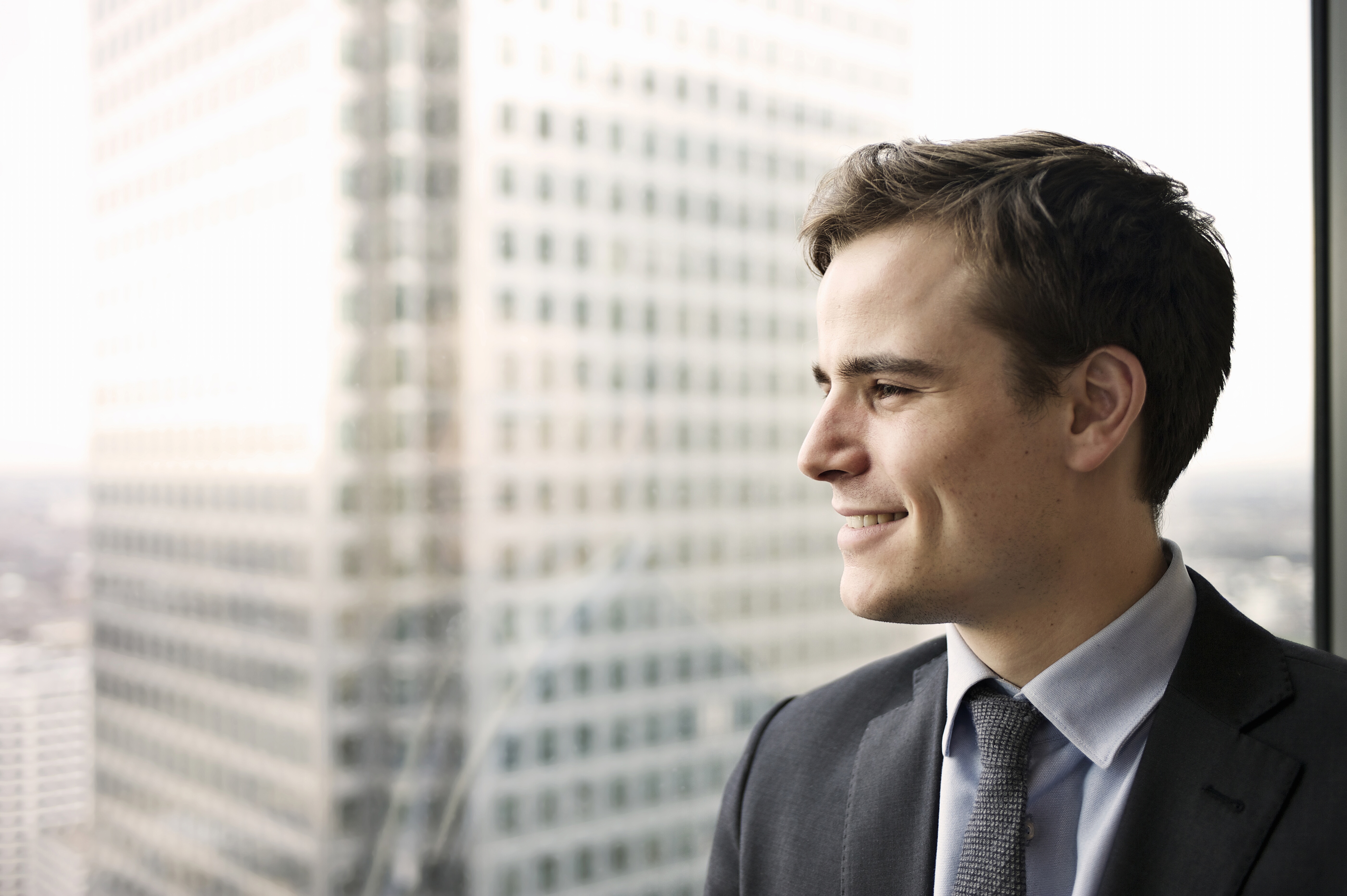 Portrait of a young business man smiling and looking out of a large window he is standing next to Portrait of a young business man smiling and looking out of a large window he is standing next to