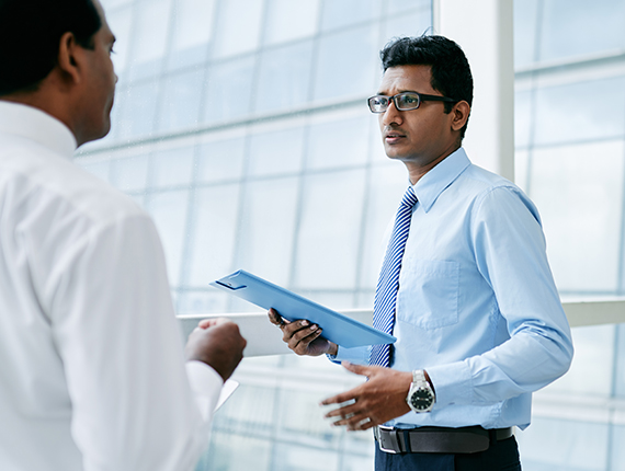 Head to waist shot of two Asian men standing in a bright room talking. The image shows the front of the man on right of picture in a blue shirt and striped blue tie holding a blue document holder with only a side view of the man on the left from his back in the frame Head to waist shot of two Asian men standing in a bright room talking. The image shows the front of the man on right of picture in a blue shirt and striped blue tie holding a blue document holder with only a side view of the man on the left from his back in the frame